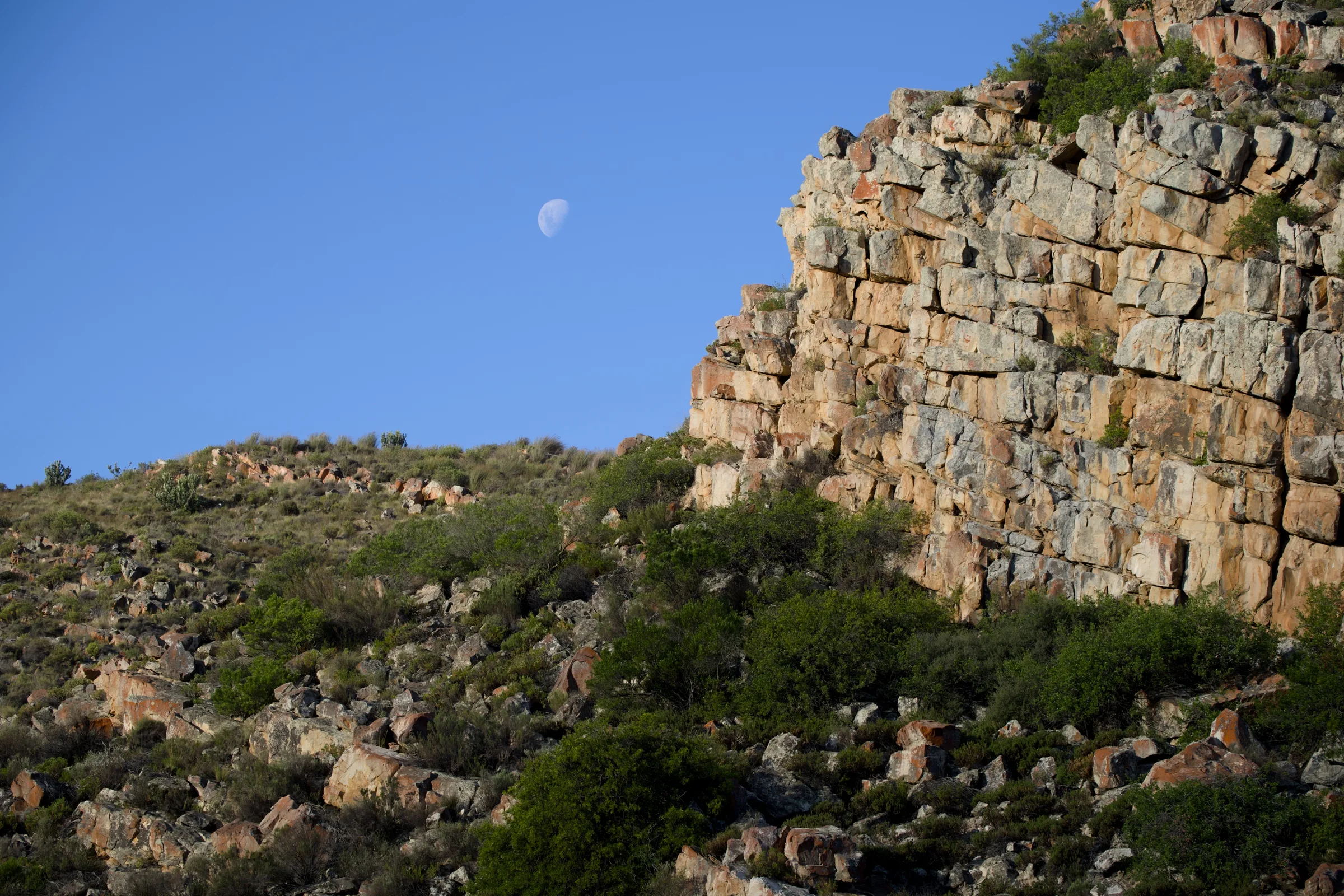 Rocky cliff face with the moon rising against a clear blue sky, AfriCamps Karoo